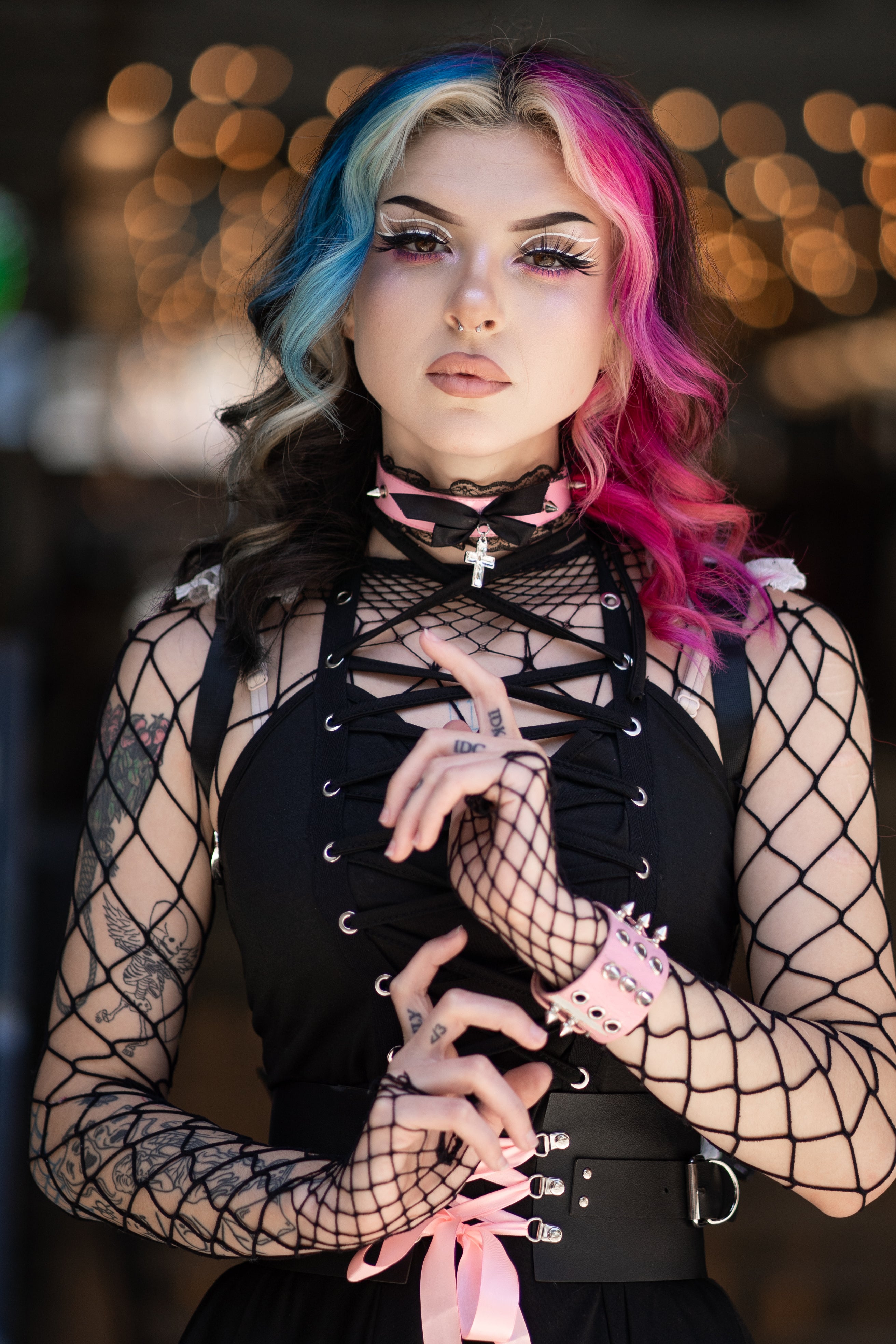 Person wearing a black lace-up dress with fishnet sleeves, colorful hair, and bold makeup, posing in a stylish setting.