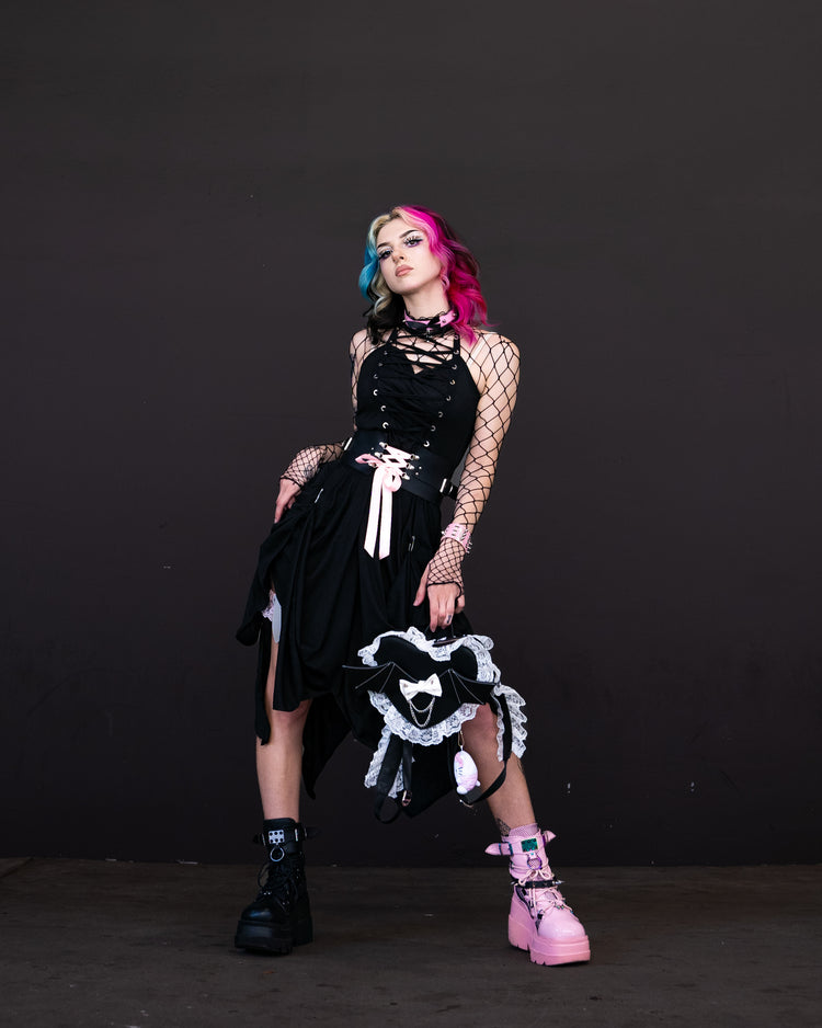 Model in colorful hair wearing a black gothic dress with lace details, fishnet sleeves, and platform boots against a dark background.