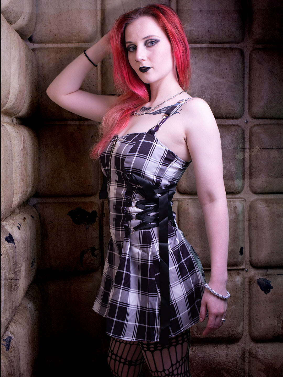 Woman in a black plaid lace-up dress with red hair, posing stylishly against a textured wall, wearing a gothic-inspired outfit.