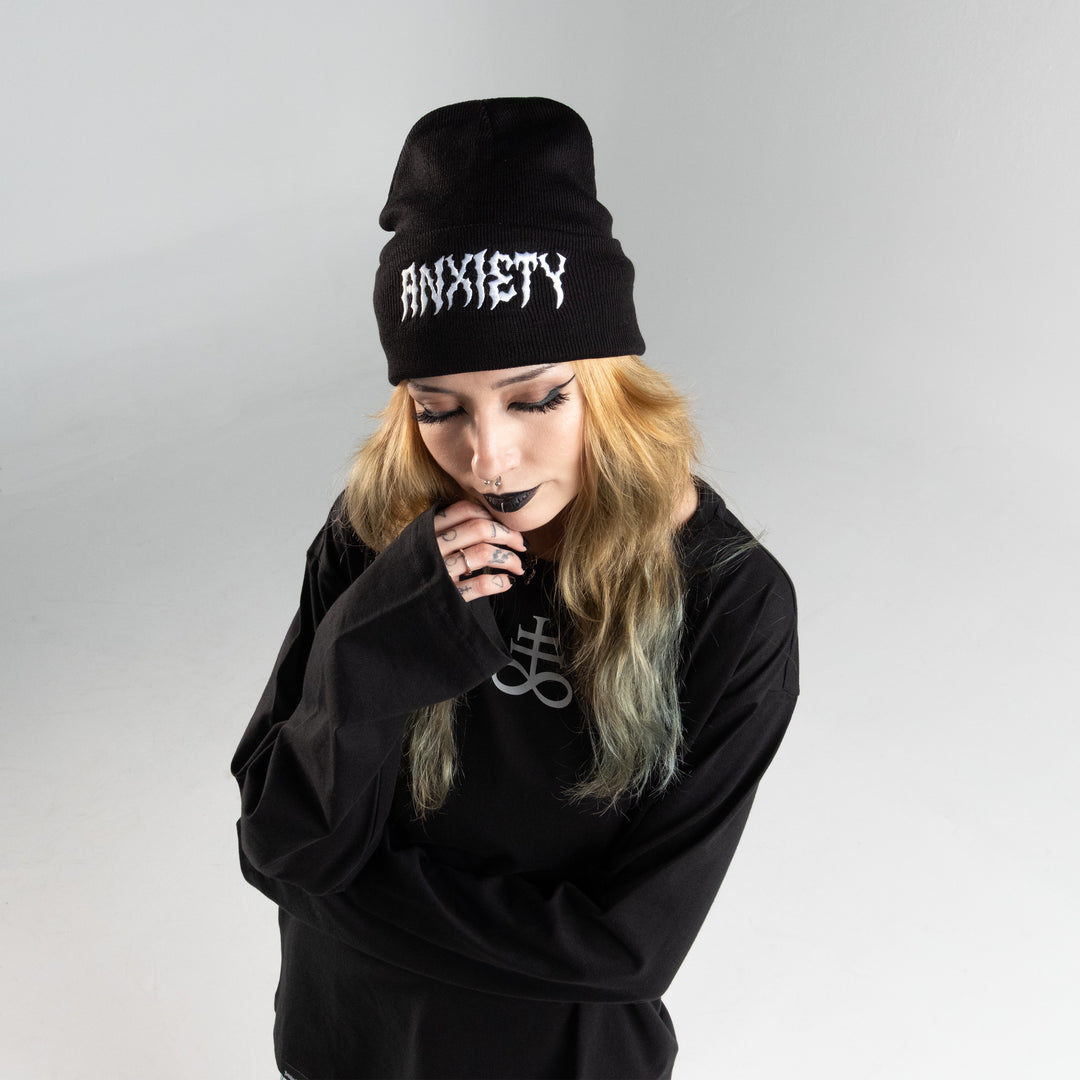 Young person wearing a black Anxiety Beanie with white embroidery, expressing a contemplative mood, part of Forbidden Alchemy collection.
